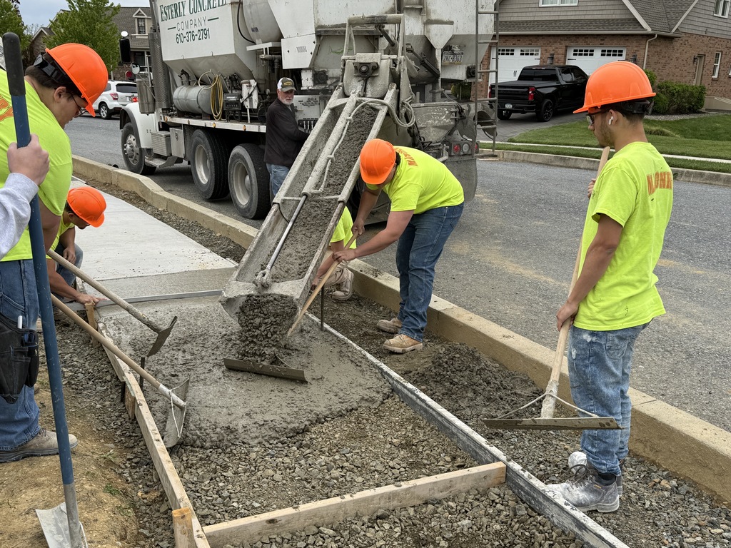 Masonry students use long-handled tools to guide and level concrete being poured from a truck into a walkway form.
