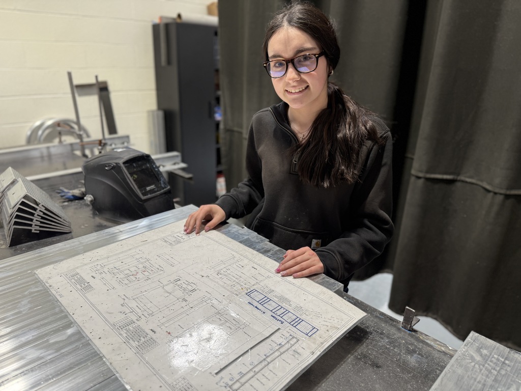 A Welding Technology student smiles while reviewing a technical blueprint on a metal work surface in a welding lab.