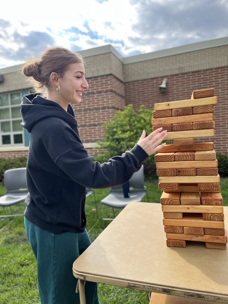 Student smiles while playing a giant wooden block stacking game outdoors.