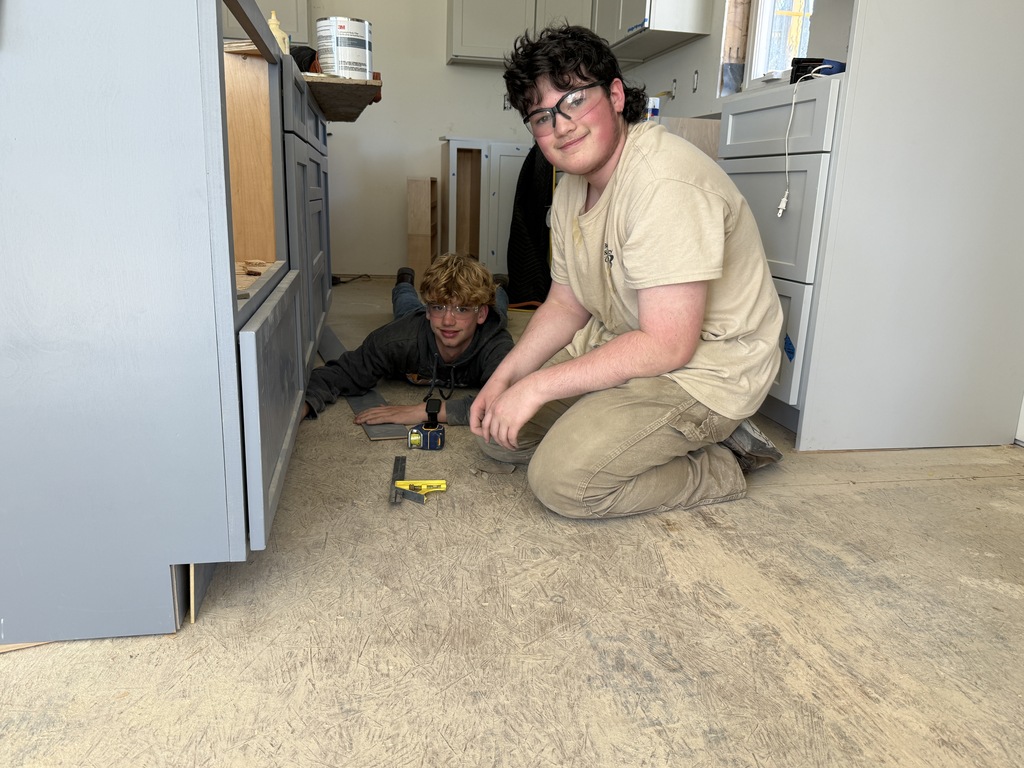 Two Cabinetry & Wood Technology students work near the base of custom kitchen cabinets inside the Student-Built House.