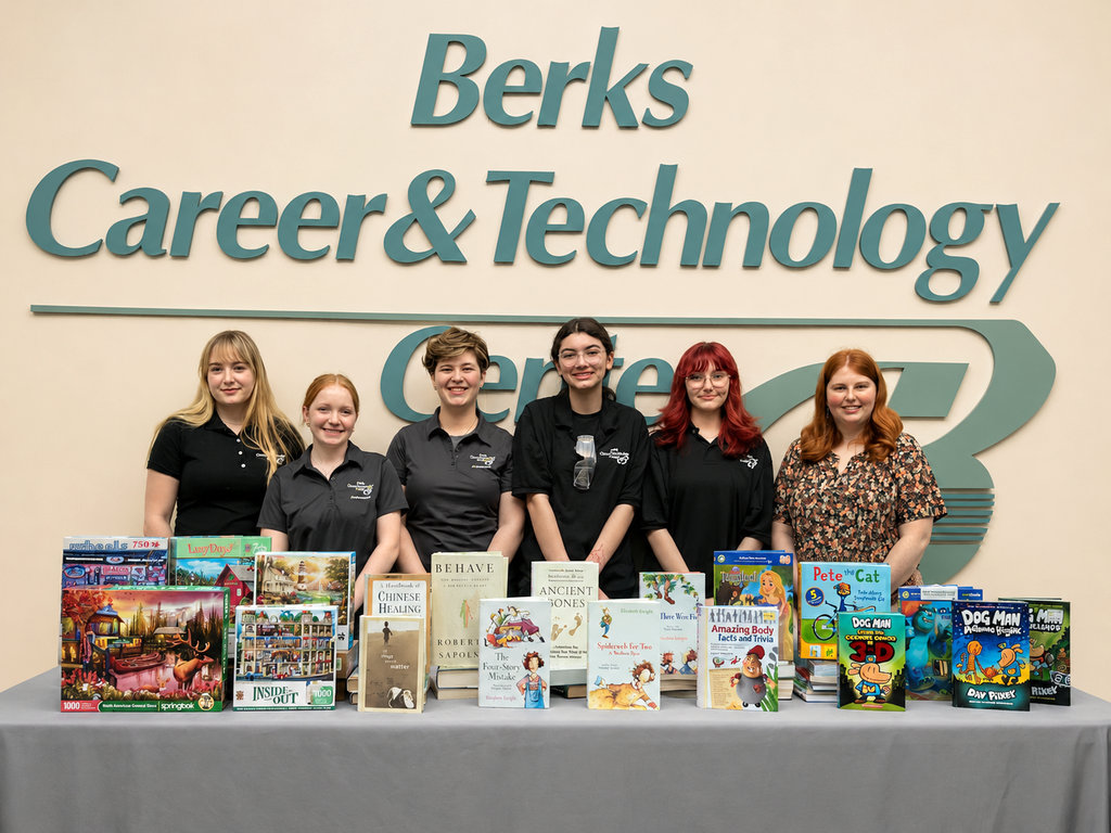 Six people stand behind a table displaying collected books, puzzles, and children’s materials for a SkillsUSA book drive at Berks Career & Technology Center.