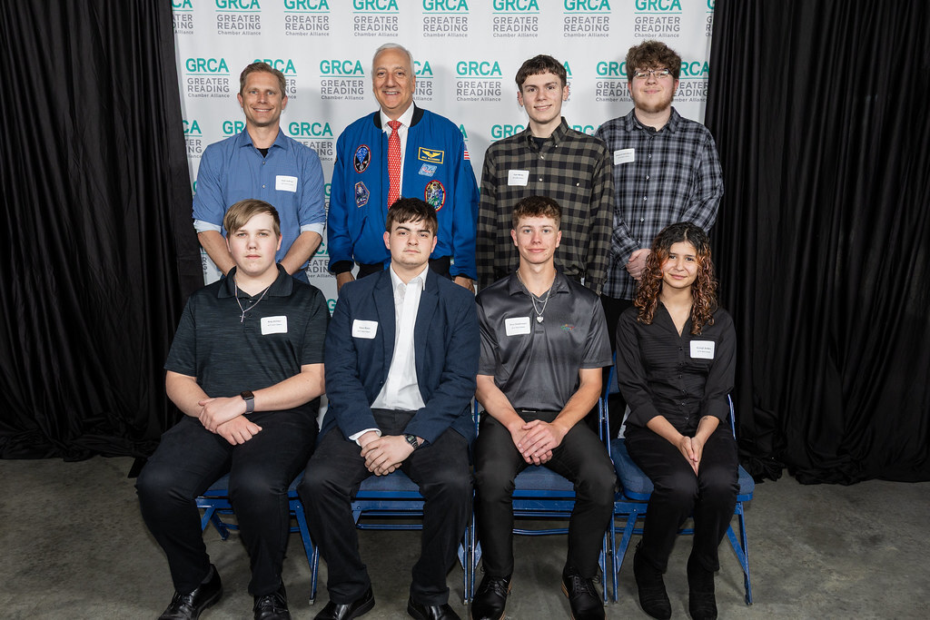 BCTC Computerized Machining Technology students pose with adults and a guest in a blue NASA flight suit in front of a Greater Reading Chamber Alliance backdrop.