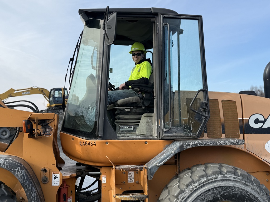 A student wearing a hard hat and safety glasses sits inside the cab of a large piece of heavy equipment outdoors.