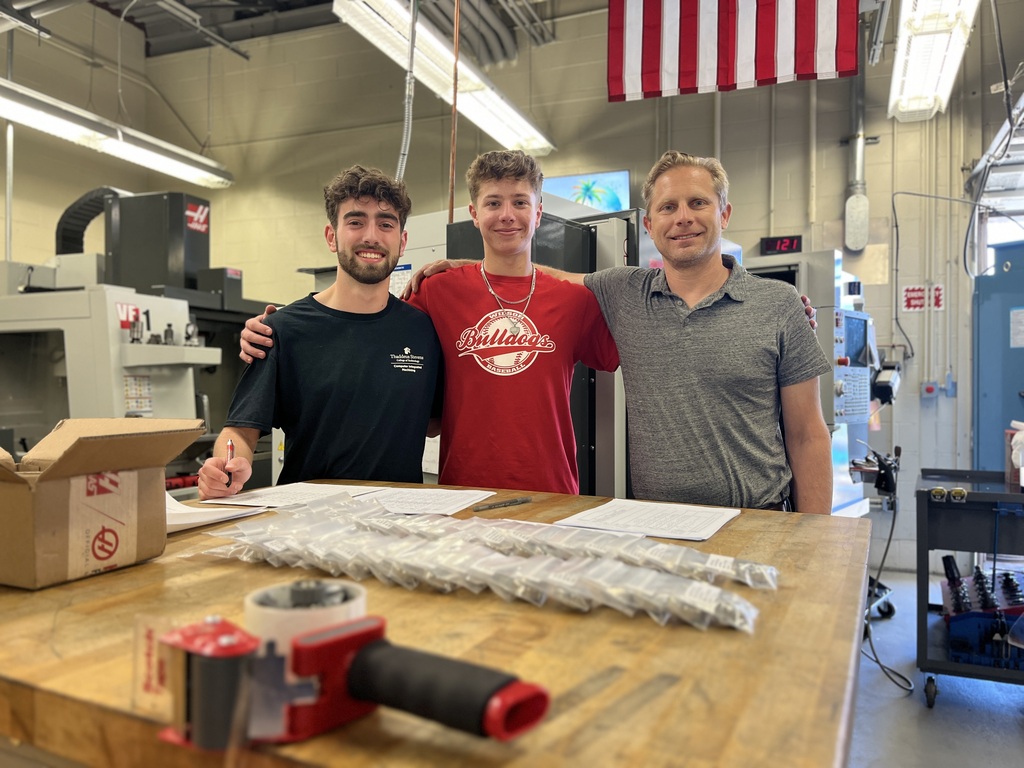 Two students and an instructor stand in a machining lab behind a table with completed NASA HUNCH project parts.