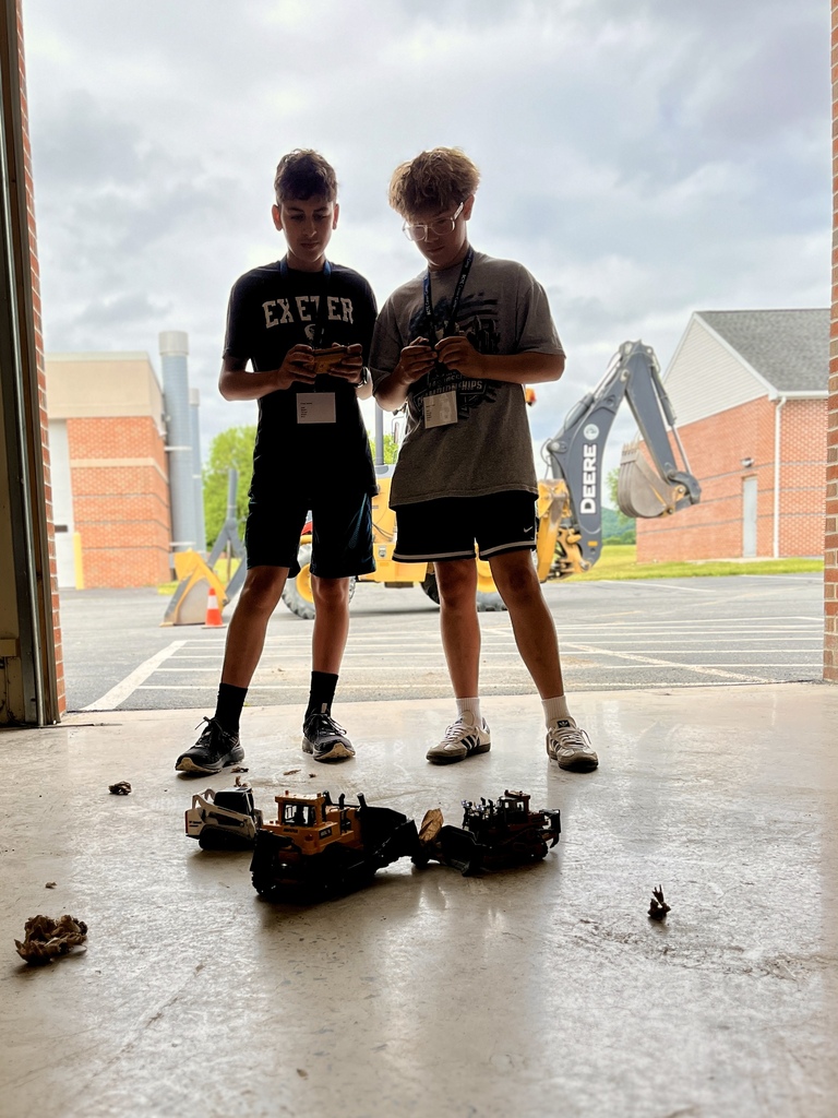 Two Career Camp participants operate remote-controlled construction vehicles near a garage bay.