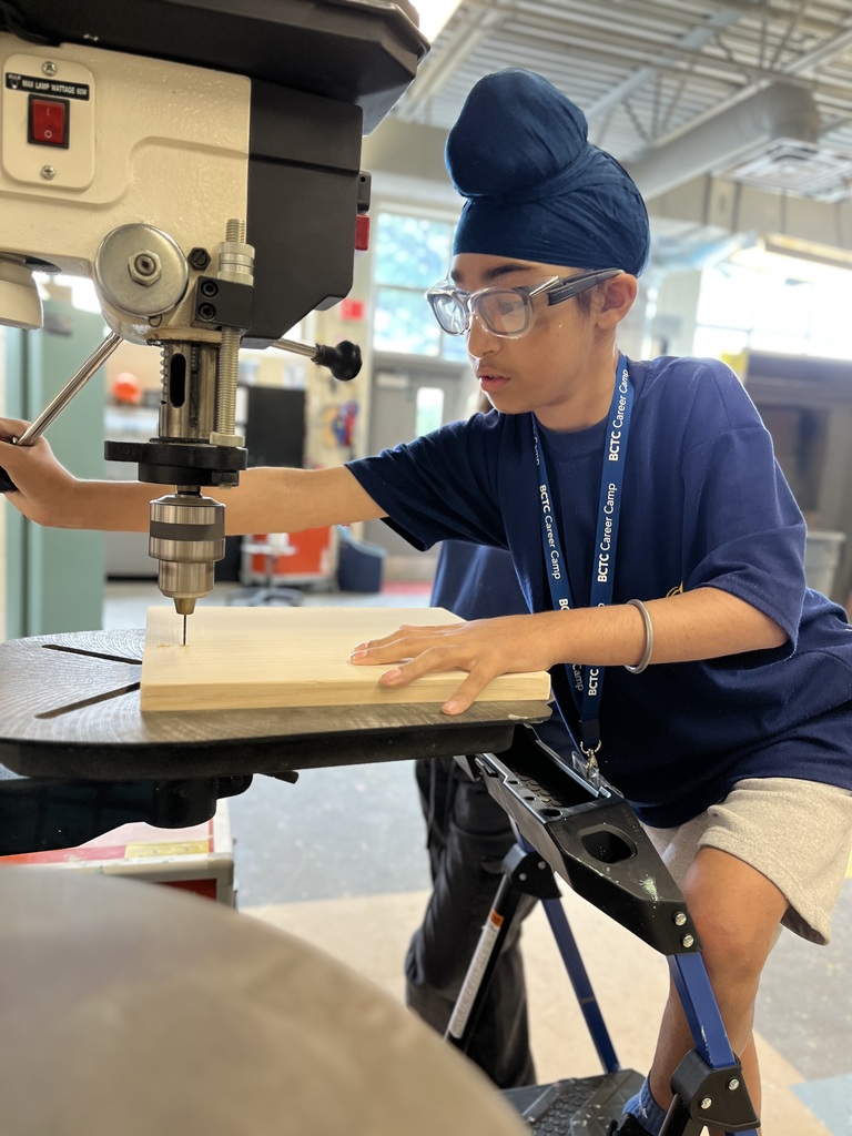 A Career Camp participant uses a drill press to make a hole in a wooden board while wearing safety glasses.
