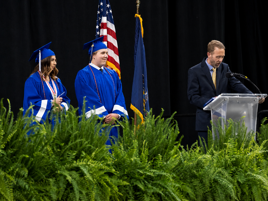 Two graduates in blue caps and gowns stand on stage during BCTC’s Senior Recognition Ceremony as a district representative speaks at a podium. American and Pennsylvania flags are displayed behind them, with ferns lining the front of the stage.