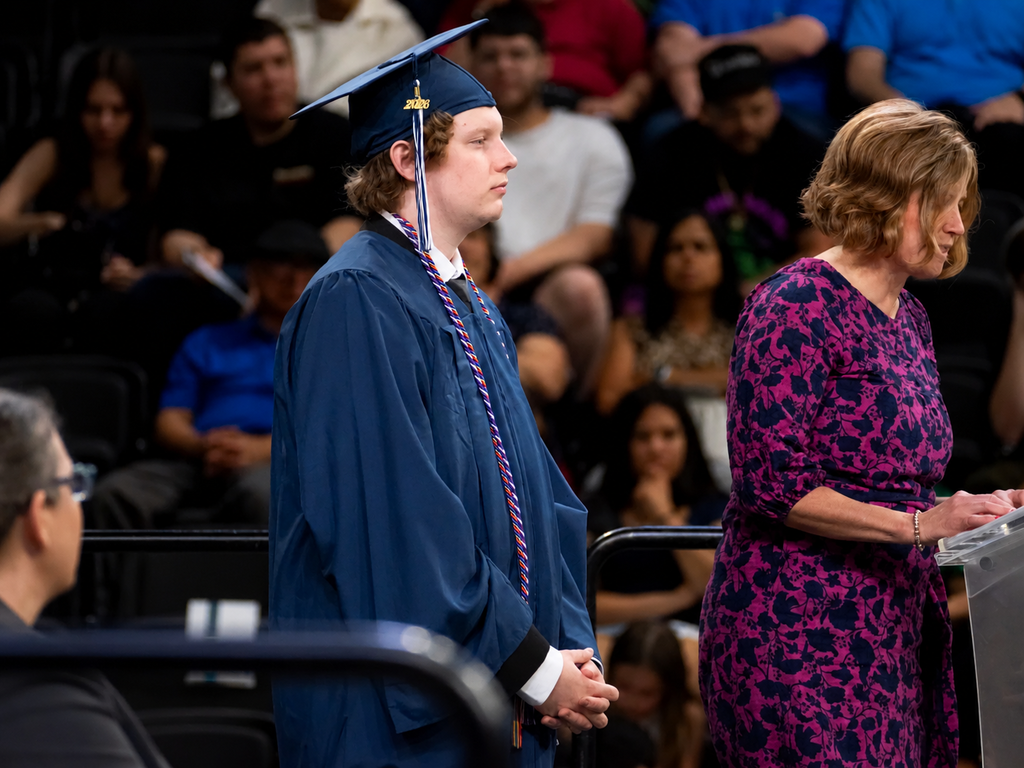 Graduate in a navy blue cap and gown stands on stage during BCTC’s Senior Recognition Ceremony while a district representative speaks at a podium. Audience members are seated in the background.