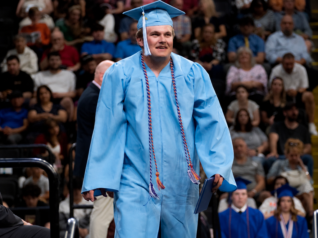 Graduate in a light blue cap and gown walks during BCTC’s Senior Recognition Ceremony while holding a diploma cover. Audience members and other graduates are seated in the background.