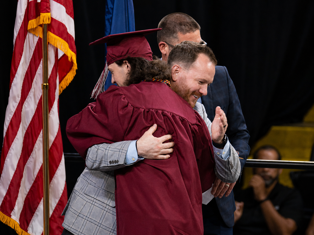 Graduate in a maroon cap and gown hugs a district representative on stage during BCTC’s Senior Recognition Ceremony. An American flag stands nearby as others look on.
