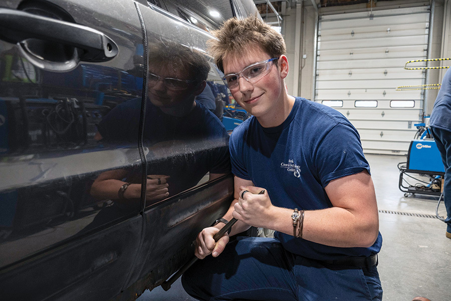 wen, an Automotive Collision Repair Technology student, kneels beside a dark-colored vehicle while working in the collision repair lab. He is wearing safety glasses and holding a hand tool near the lower side panel of the vehicle.