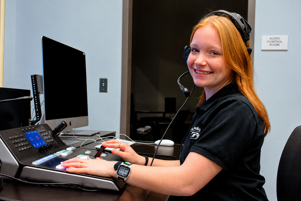 Samantha, a Video & Media Content Production student, smiles while wearing a headset and working at an audio control console in a media production room.