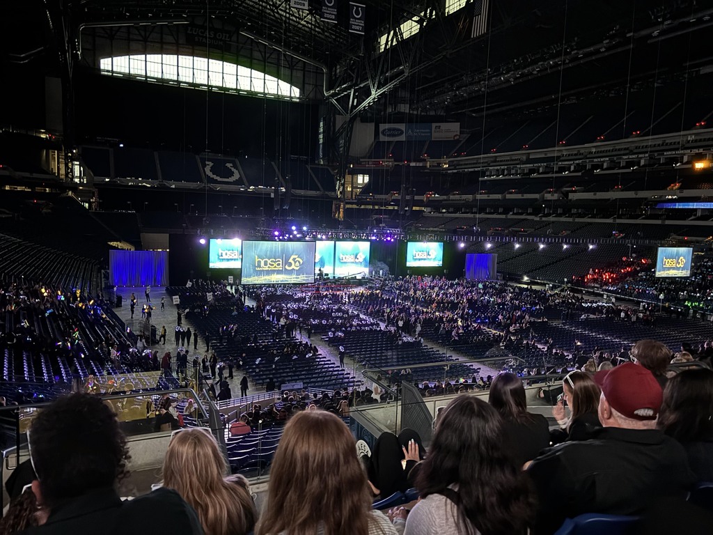 A large crowd attends a HOSA International Leadership Conference session inside Lucas Oil Stadium, with the stage and presentation screens visible.