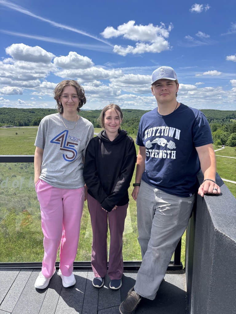 Three BCTC HOSA students stand together on an outdoor overlook with green hills, trees, and a blue sky with clouds behind them.