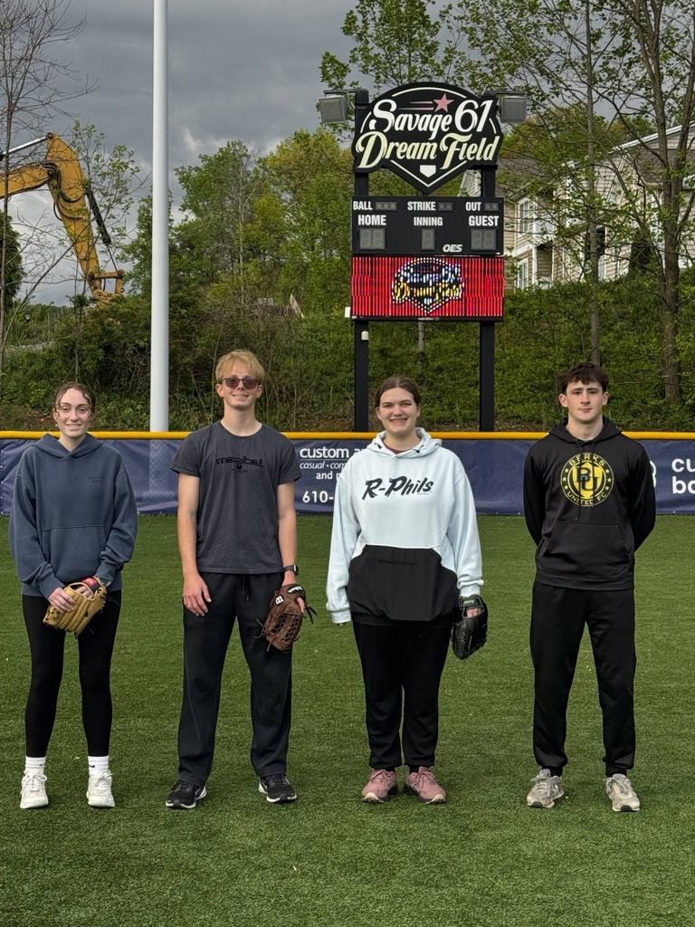 Four BCTC students stand on the turf at Savage 61 Dream Field, holding baseball gloves in front of the field’s scoreboard.