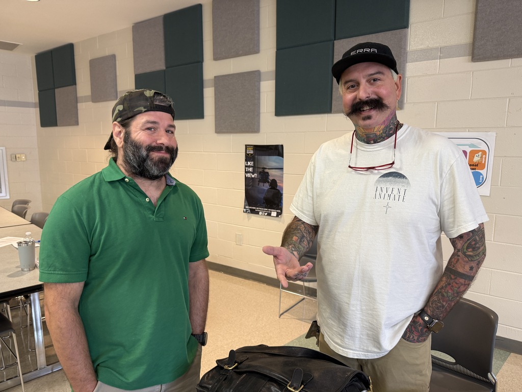 Two men in a room with a white wall, grey panels, and chairs. One man has a beard and wears a green shirt. The other has a mustache, wears a white shirt, and has tattoos.