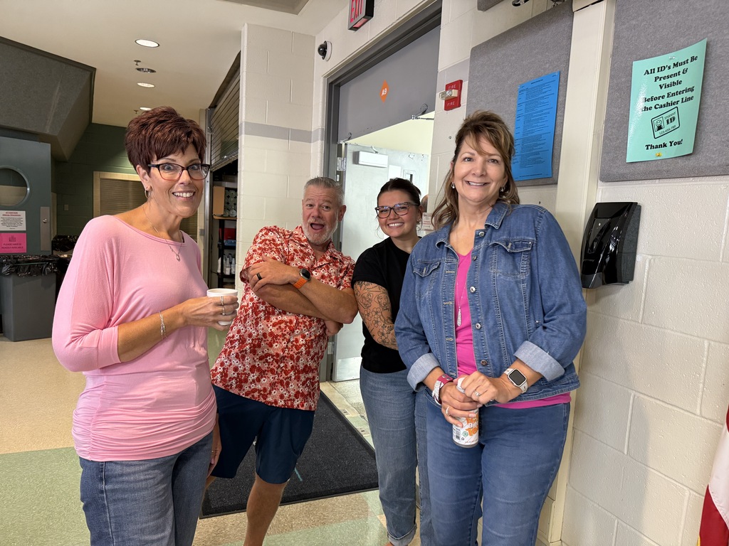Four adults stand in a hallway, with two women holding drinks and two men standing behind. A trash can and a sign are in the background.