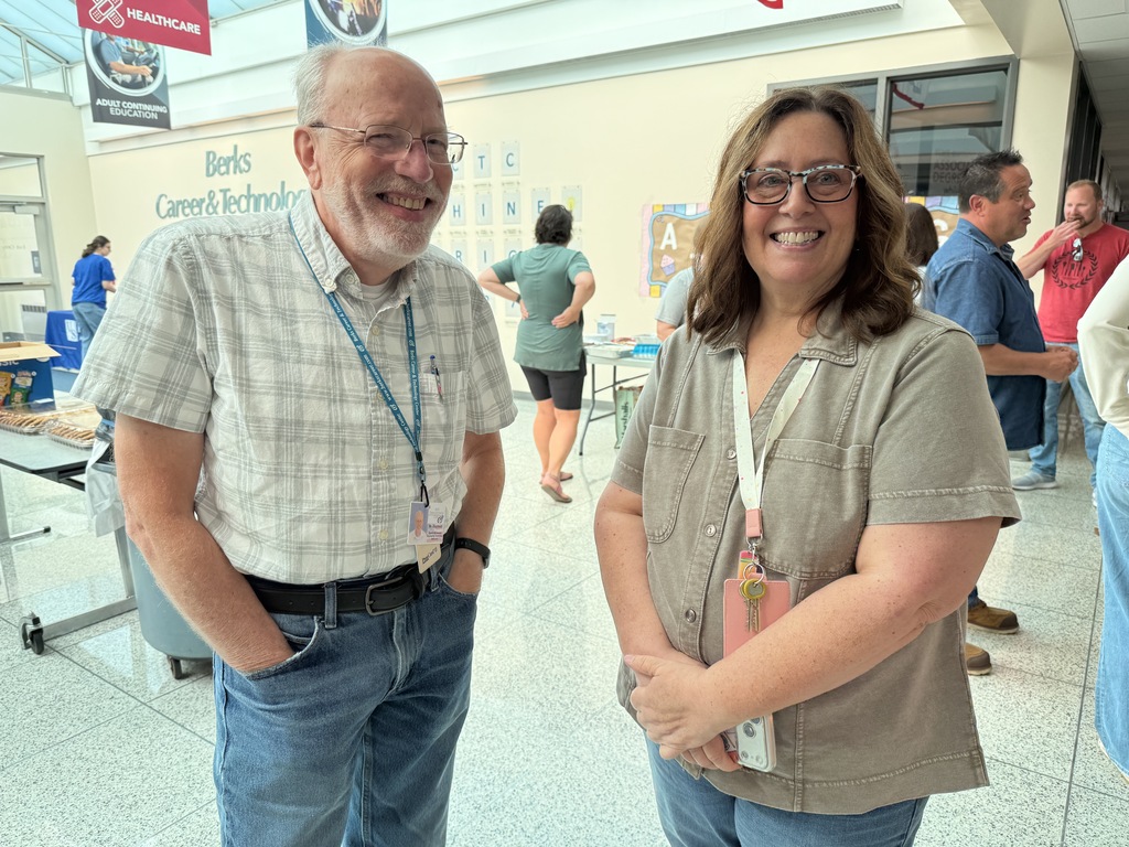 A man and a woman smile for a photo. They stand in a brightly lit hallway.