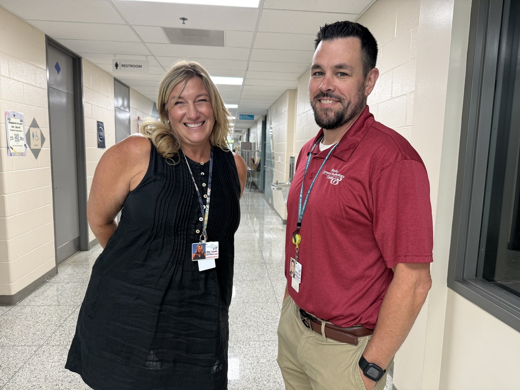 A man and a woman stand in a hallway with white walls and tile floors, both smiling.