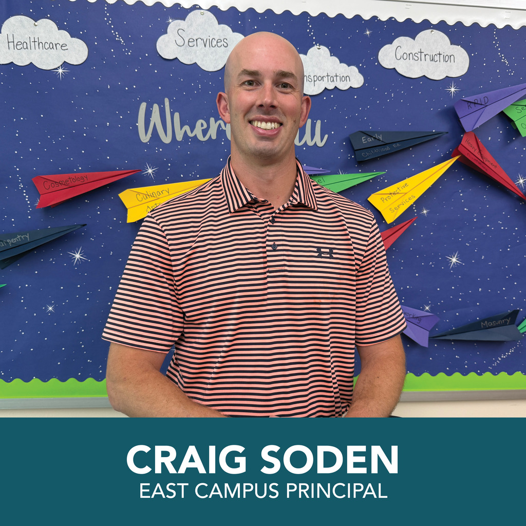 A man with a bald head stands smiling in front of a bulletin board with paper airplanes.