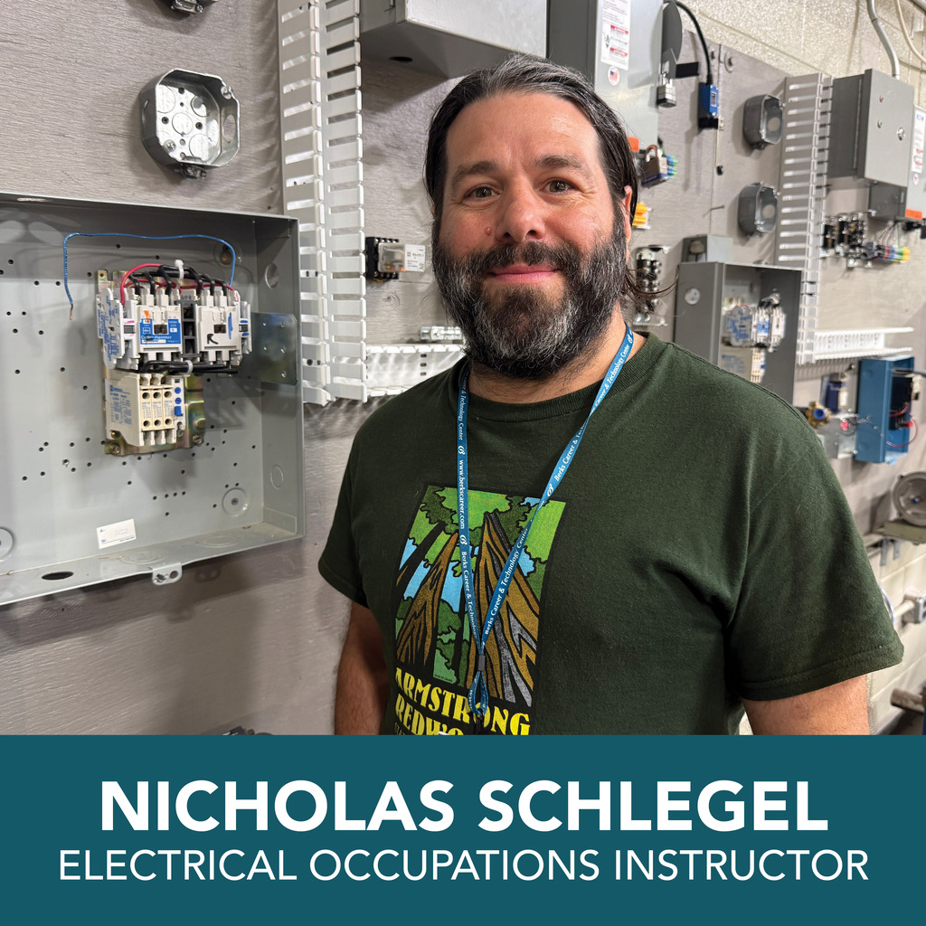A man in a green shirt, with a beard, stands in front of electrical equipment. A blue lanyard hangs around his neck.