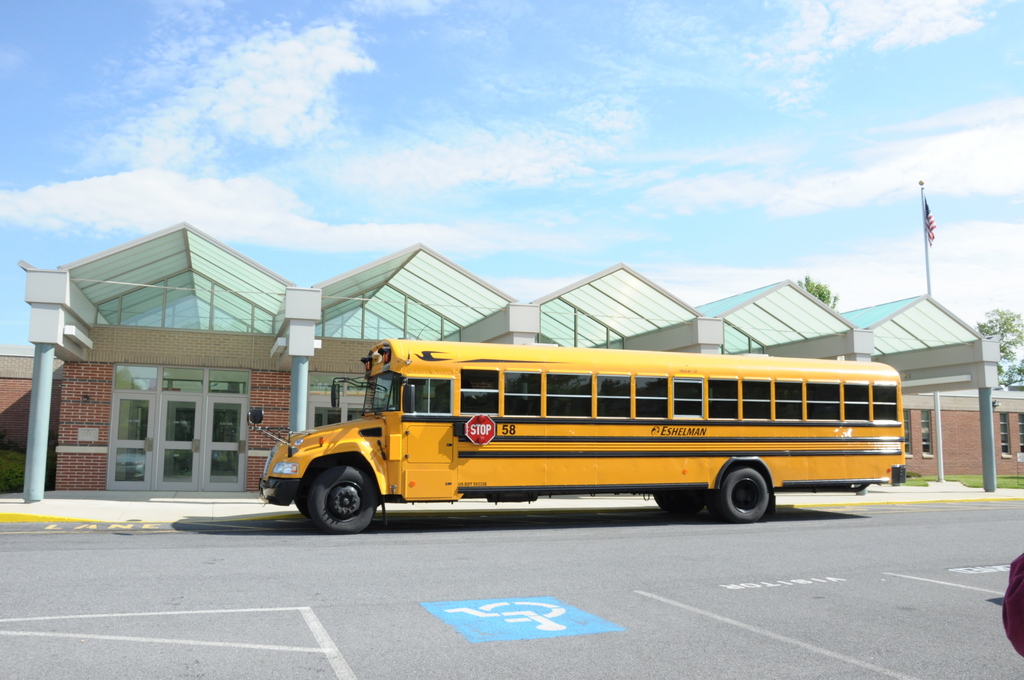 A yellow school bus is parked in front of a building with glass roofs. The sky above is blue with some clouds.