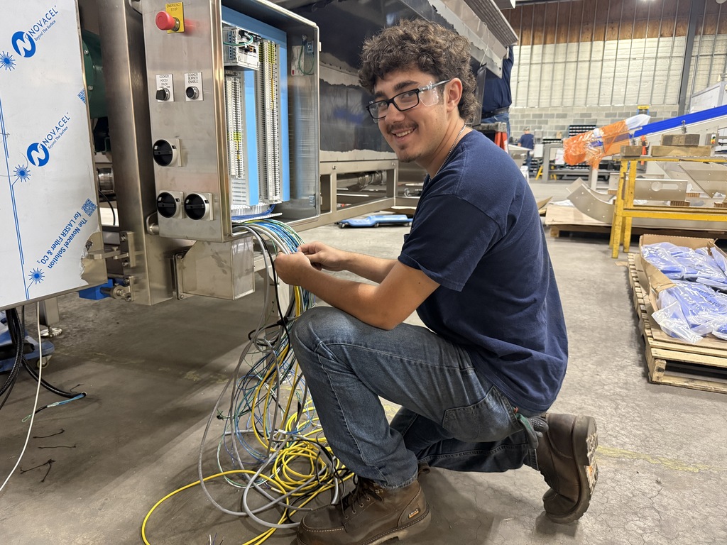 A man in a blue shirt and glasses is kneeling in a factory, working on wires.