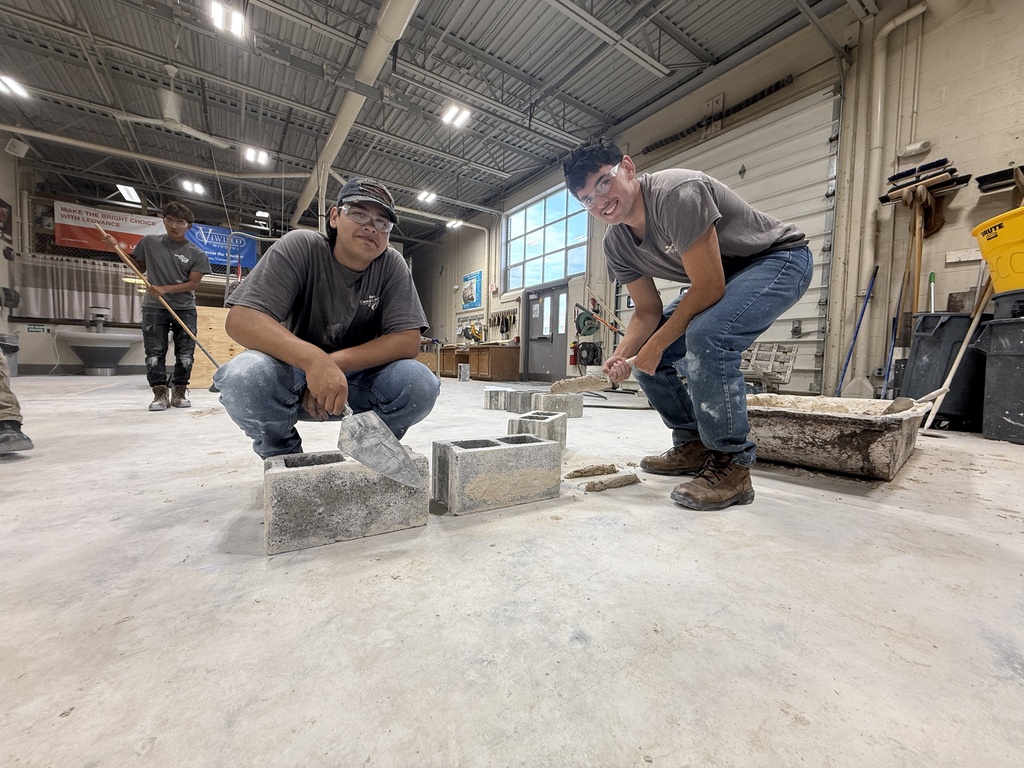Two men work in a warehouse, one kneeling with tools and the other standing, both wearing hats and jeans.