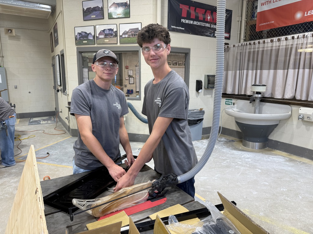 Two men in gray t-shirts and glasses work together at a table in a workshop. Tools and materials are scattered around them.