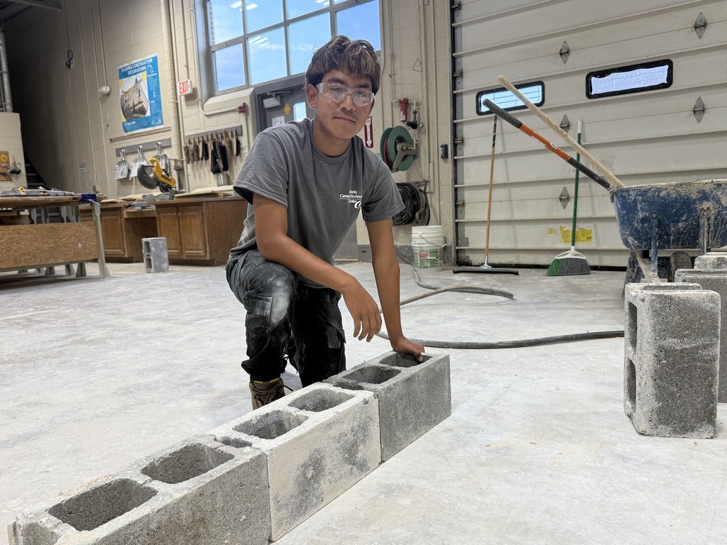 A young person in a gray shirt and glasses works with cinder blocks on a concrete floor.