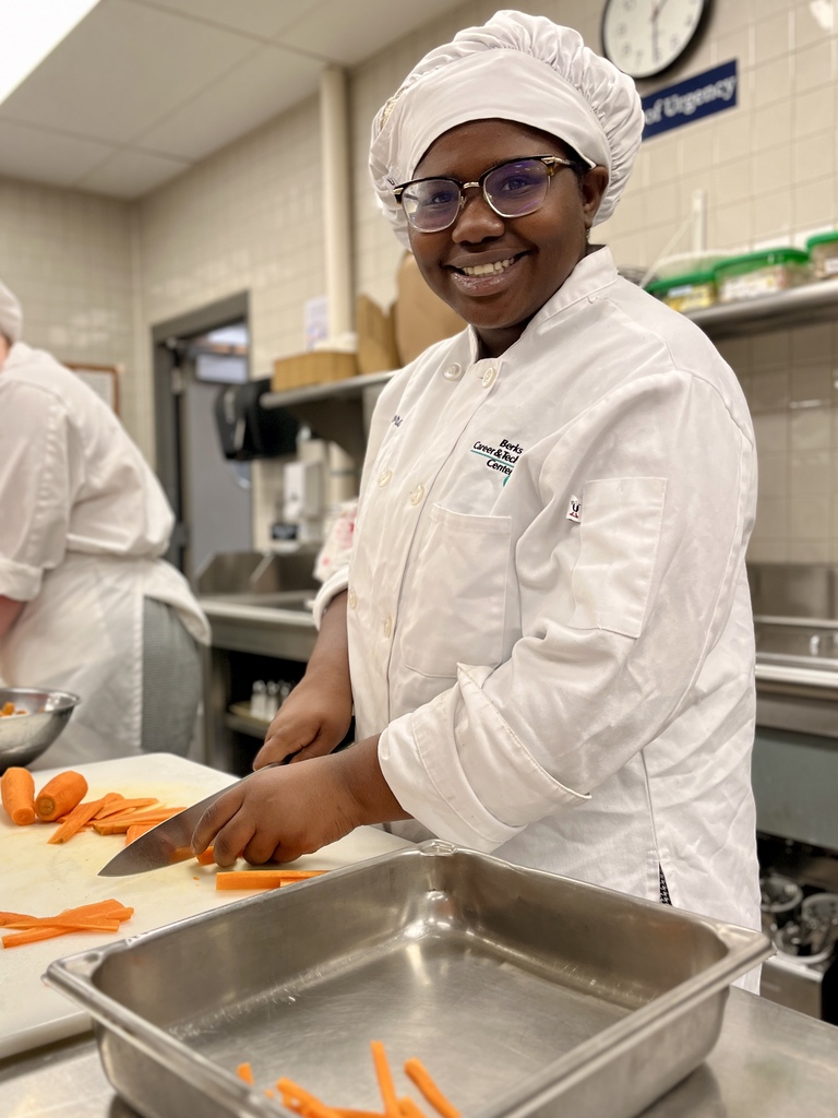 Person in chef's uniform chops carrots on a cutting board in a kitchen. Another person works in background.