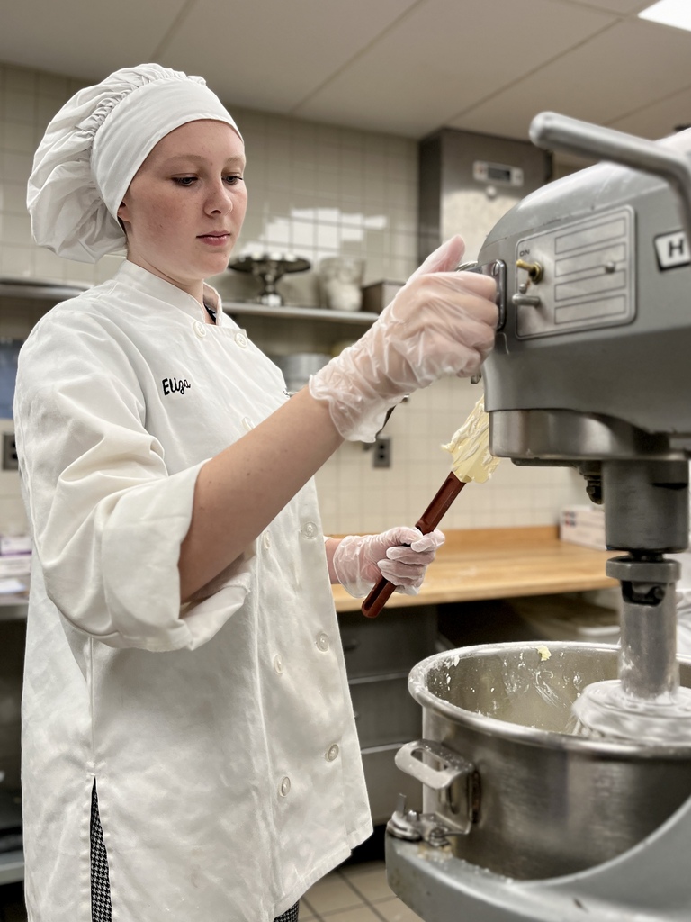 Person in chef's uniform holding a whisk and working with a large stand mixer.