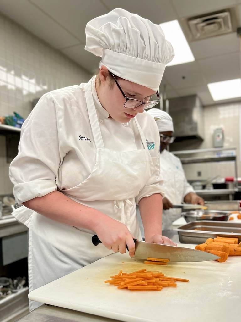 Person in a white chef's uniform with a hat and glasses cutting carrots on a white cutting board.