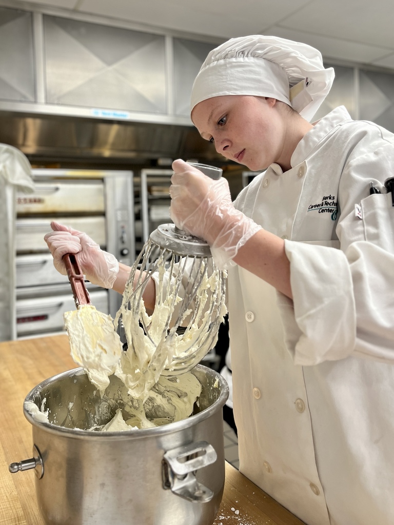 A chef in a white uniform, hat, and gloves uses a mixer to blend ingredients in a bowl.
