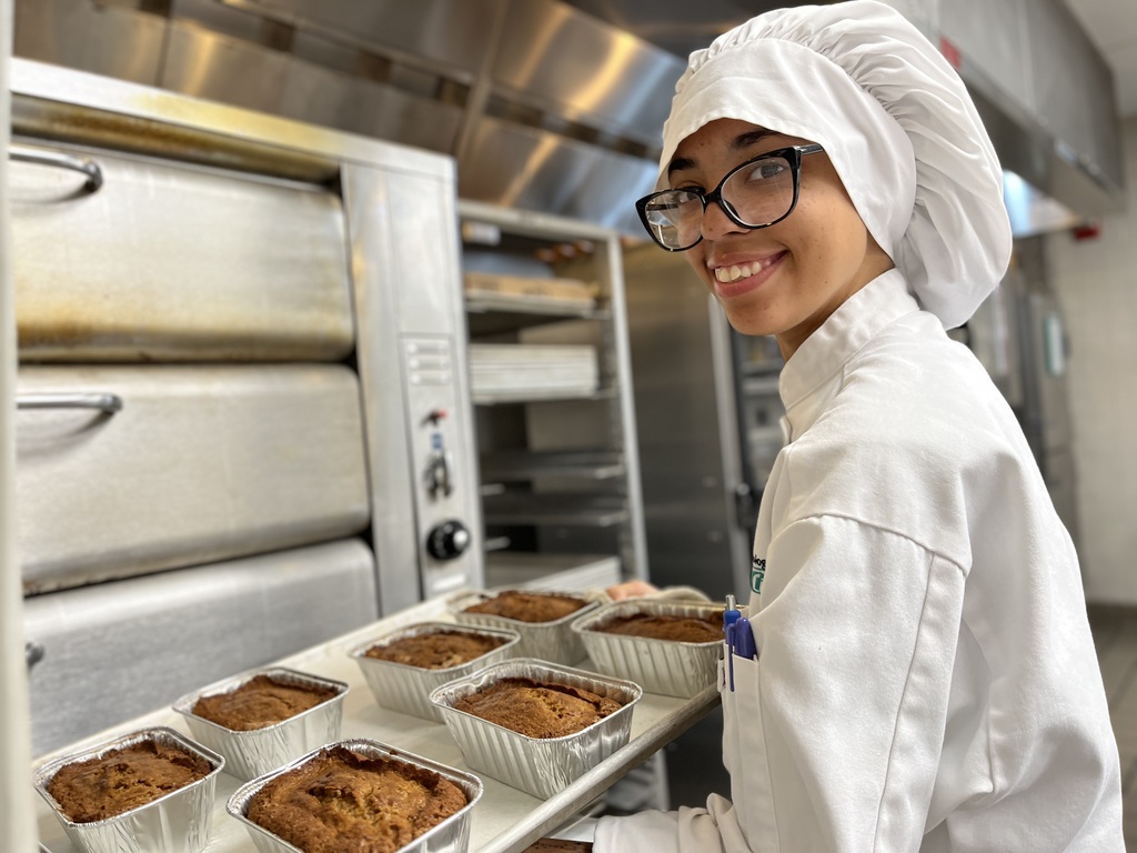 Person in chef's outfit holding a tray with baked goods in a kitchen with industrial oven.