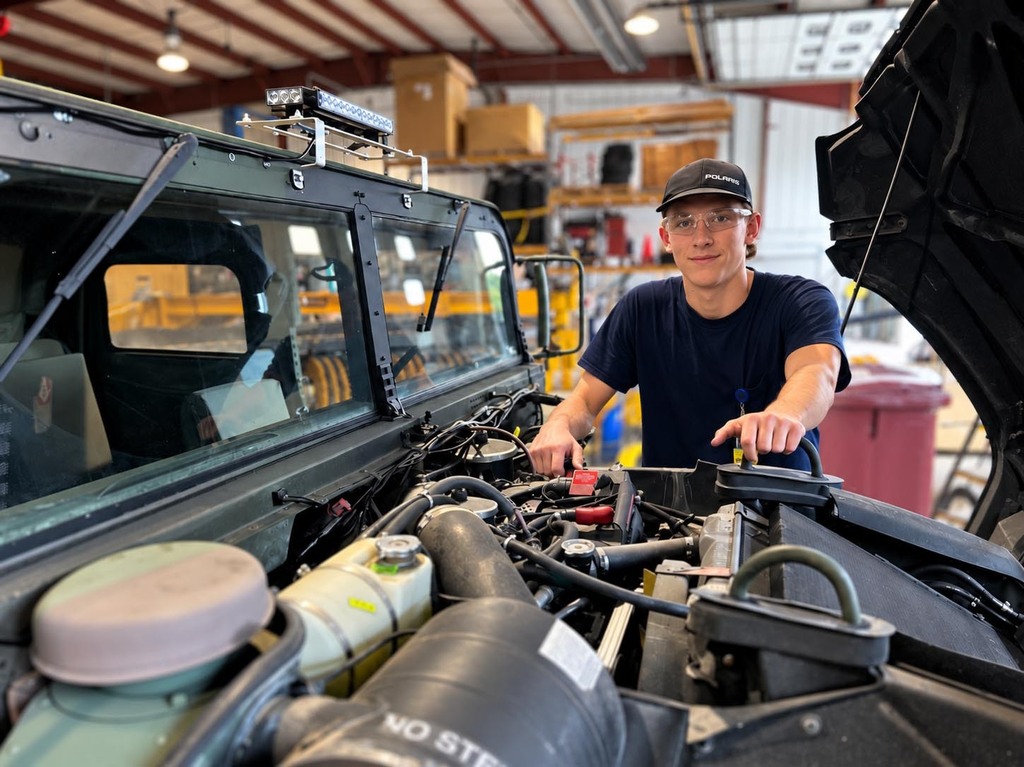 A man in a workshop with a hood up, working on a vehicle's engine.