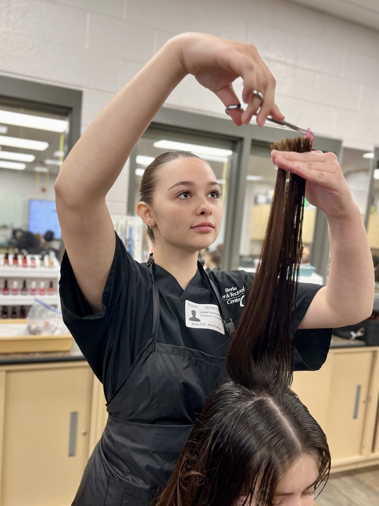 A woman in a black apron cuts another woman's hair with scissors in a salon.