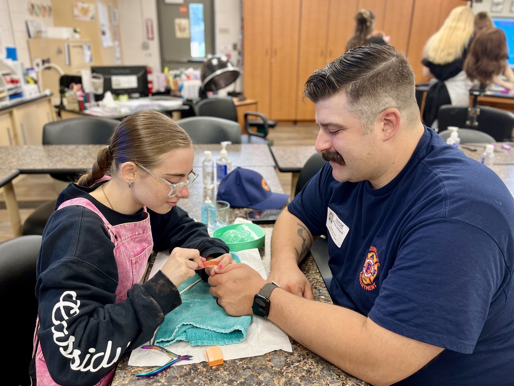 A girl wearing glasses works on a blue cloth while a man with a mustache helps her.