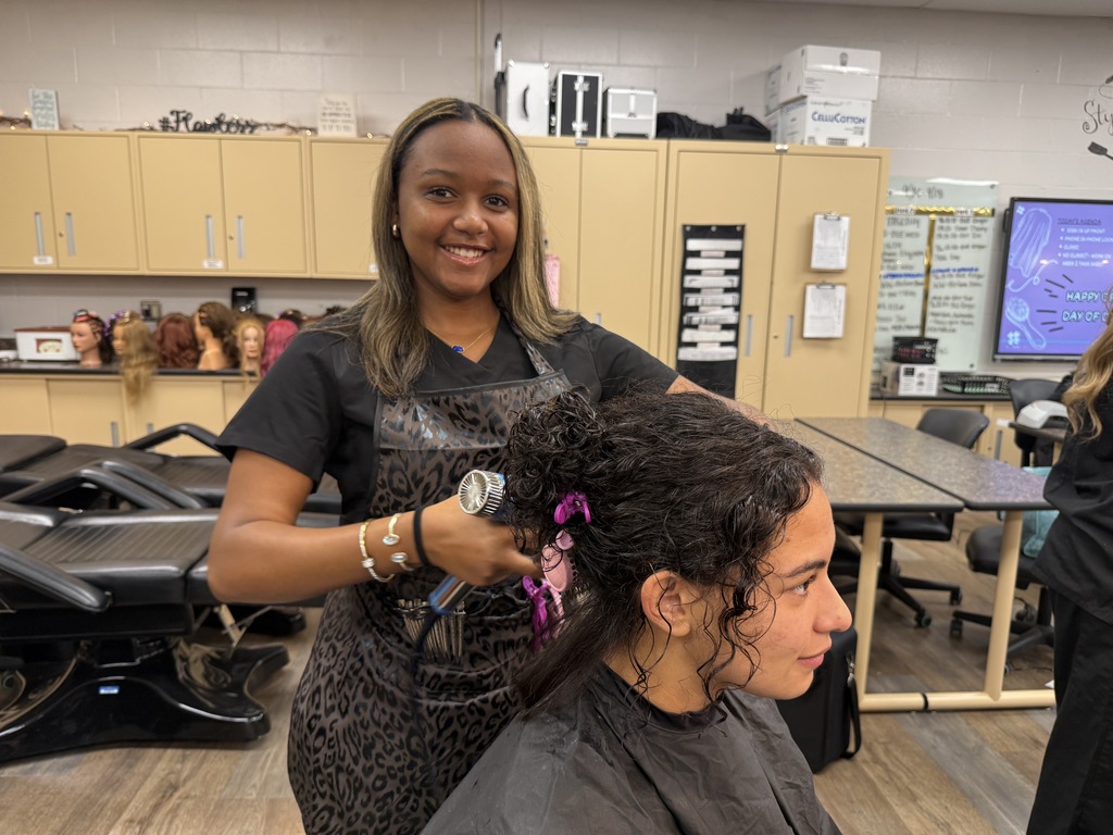 A woman in an apron stands behind another woman seated in a chair, using a tool to style her hair in a classroom.