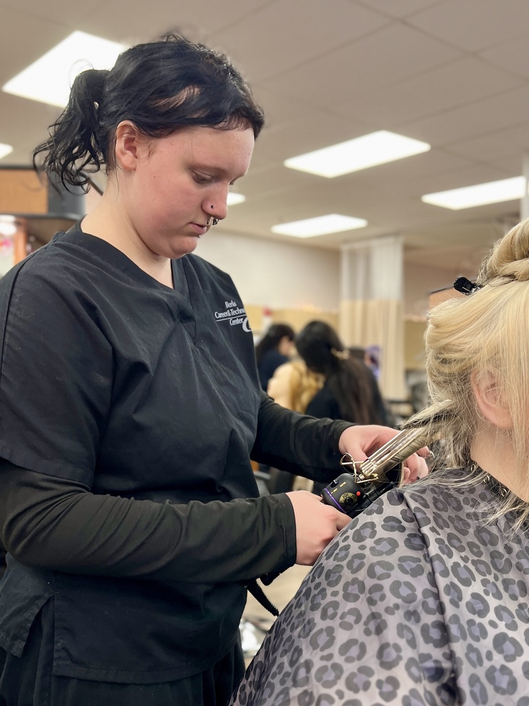 A woman is styling another woman's hair at a salon; she wears a black uniform and the client has a leopard-print cape.
