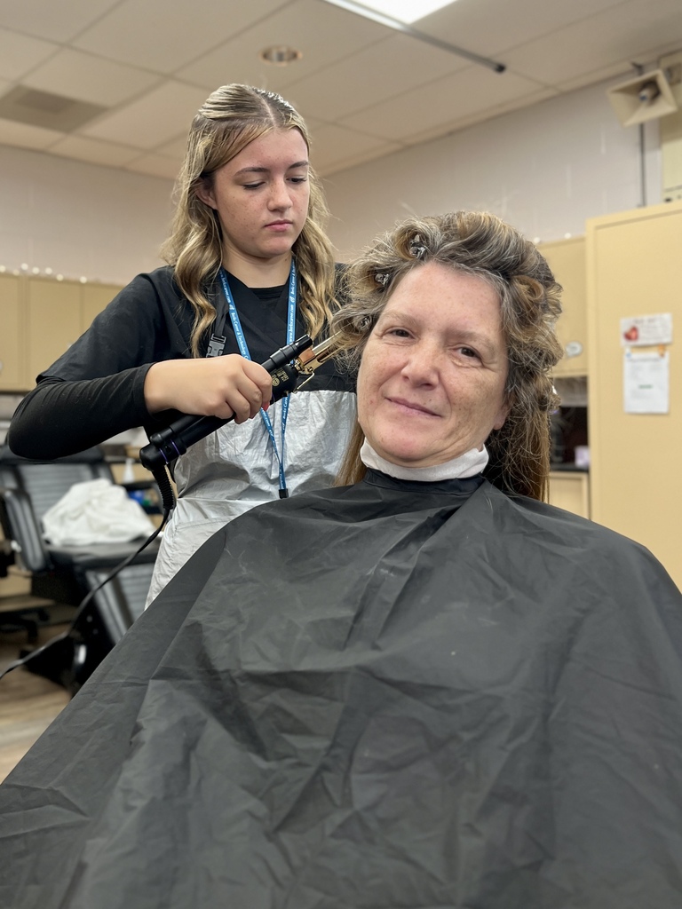 A woman sits in a salon chair as another person styles her hair with a curling iron.