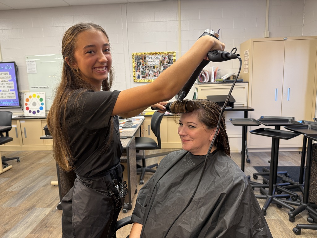 A woman uses a blow dryer to style another woman's hair in a salon, with wood floors.