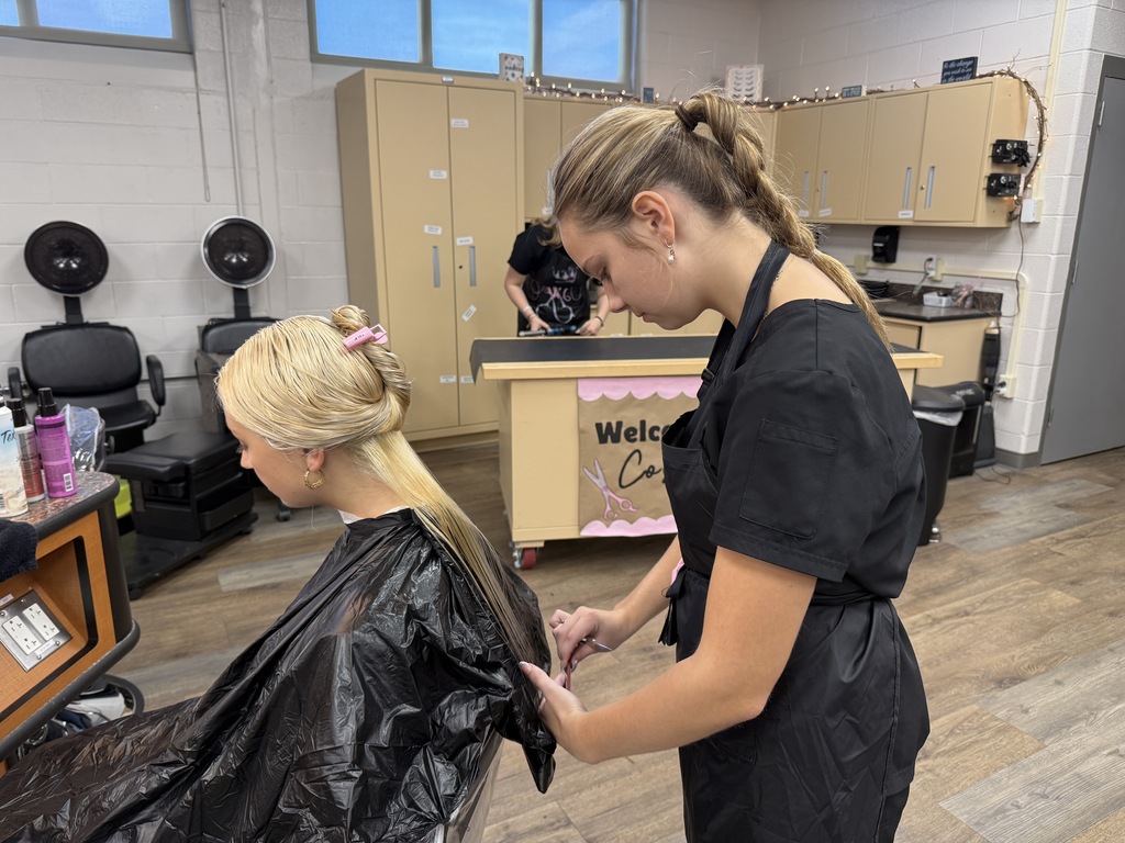 A person stands over a seated woman, cutting her hair in a salon with wooden floors.