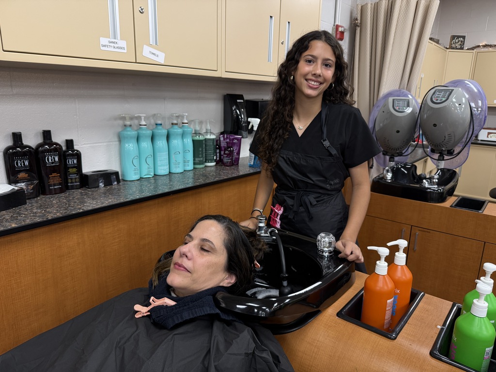 A woman in a salon chair, receiving a shampoo service from another woman, with shelves and bottles in the background.