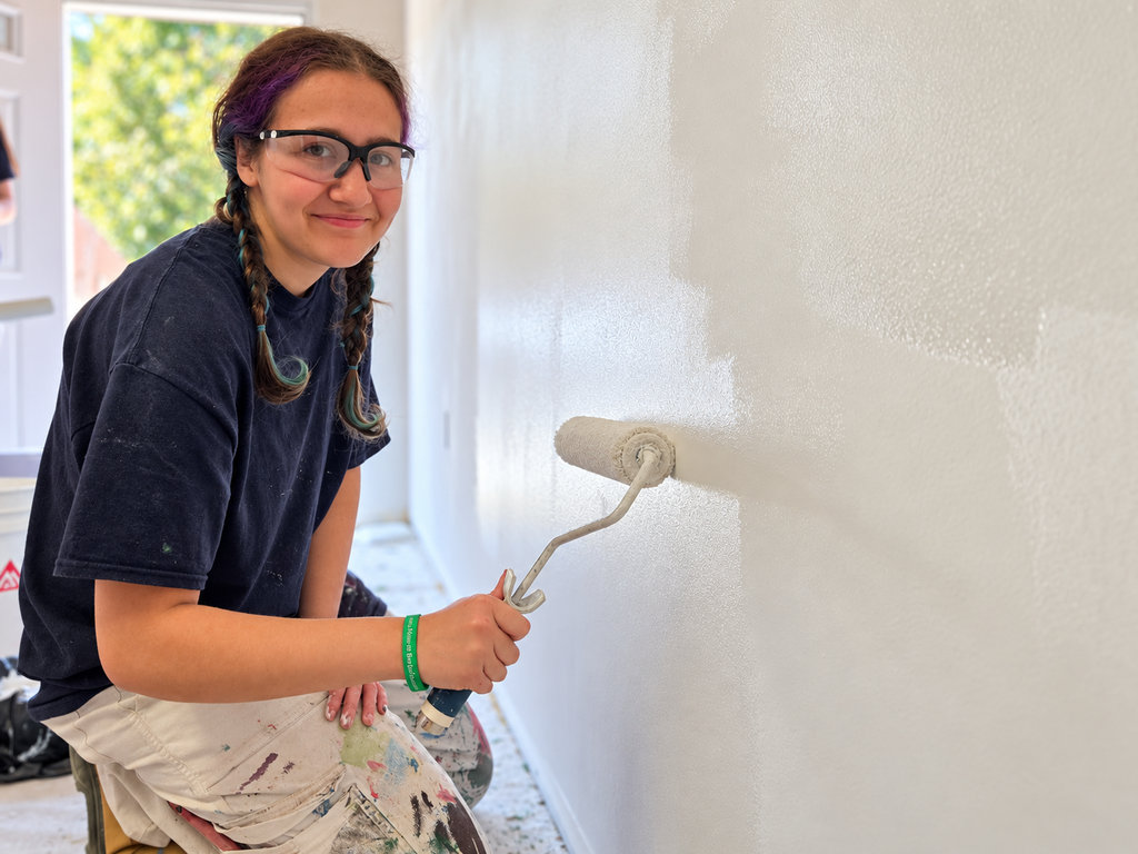 Person with glasses and braids painting a white wall using a roller. White paint on the roller.