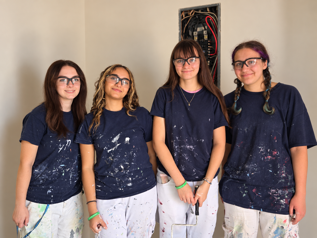 Four girls in paint-stained shirts and glasses pose for a photo in a room. A wall with a black frame is behind them.