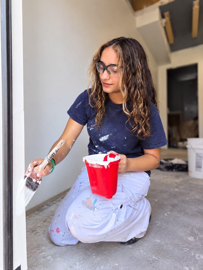 A person crouches on the floor holding a paintbrush, with a red bucket of paint.