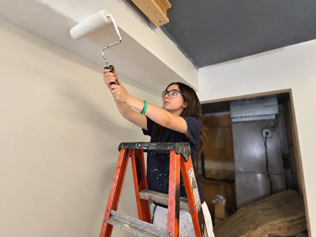 A person stands on an orange ladder, using a roller to paint a ceiling white. The wall is light brown.