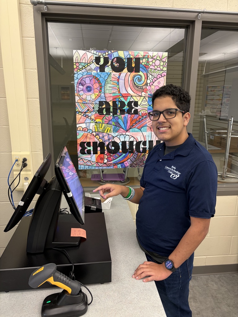 Man in a blue shirt and glasses stands behind a counter with a computer. Behind him is a window with a motivational poster.