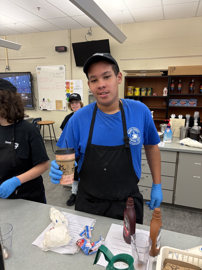 Person in blue shirt and apron holds a cup and stands at a counter with food prep items.